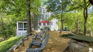 A rustic outdoor seating area with benches and a stone table, surrounded by trees and a building with a green facade. - Warm + Peaceful Home w/ Fire Pit on Haw River (Pittsboro)