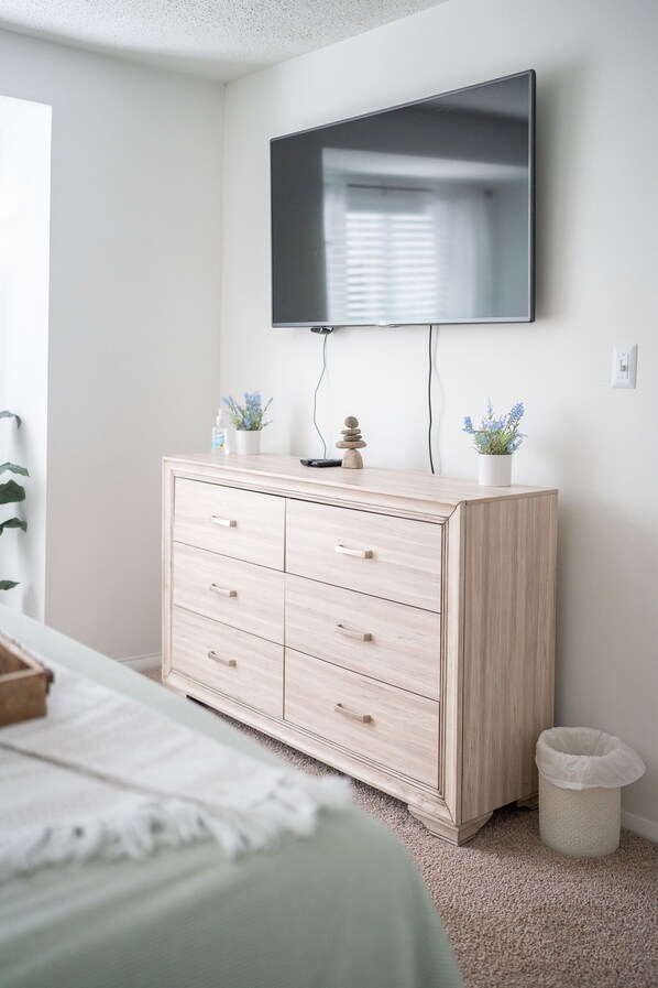 A bedroom with a bed, a wooden dresser, a television mounted on the wall, and a small trash can.
