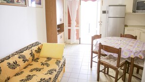 Sala de estar com sofá, mesa de jantar com cadeiras, cozinha com geladeira e porta que dá para o exterior.