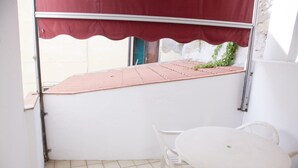 Pequeno alpendre externo com chão de azulejos, uma mesa branca e duas cadeiras brancas.