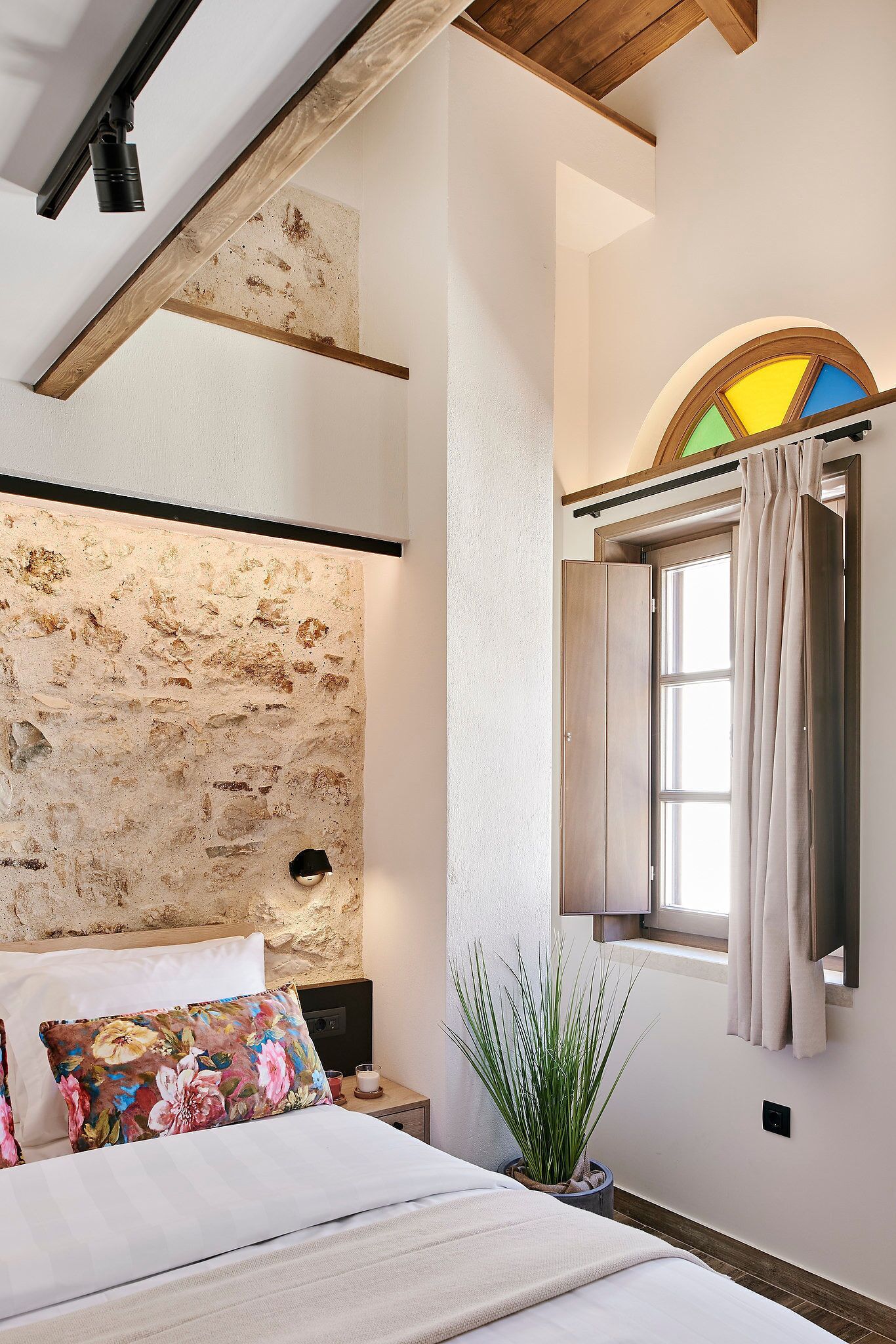 Un dormitorio con pared de piedra, cama con almohadas florales, mesita de noche con planta y ventana con cortinas.