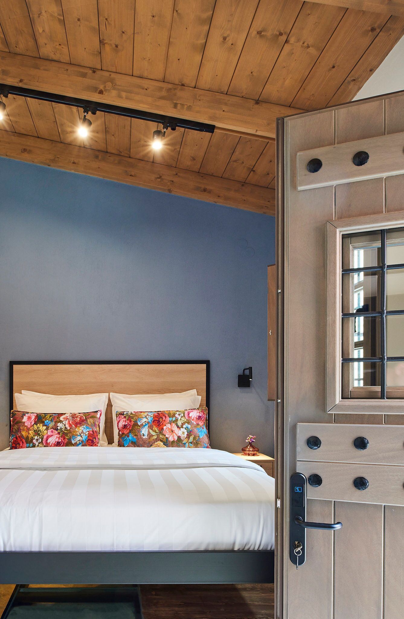 A bedroom with a wooden ceiling, a bed with white bedding and floral pillows, a nightstand with a lamp, and a wooden door with a window.