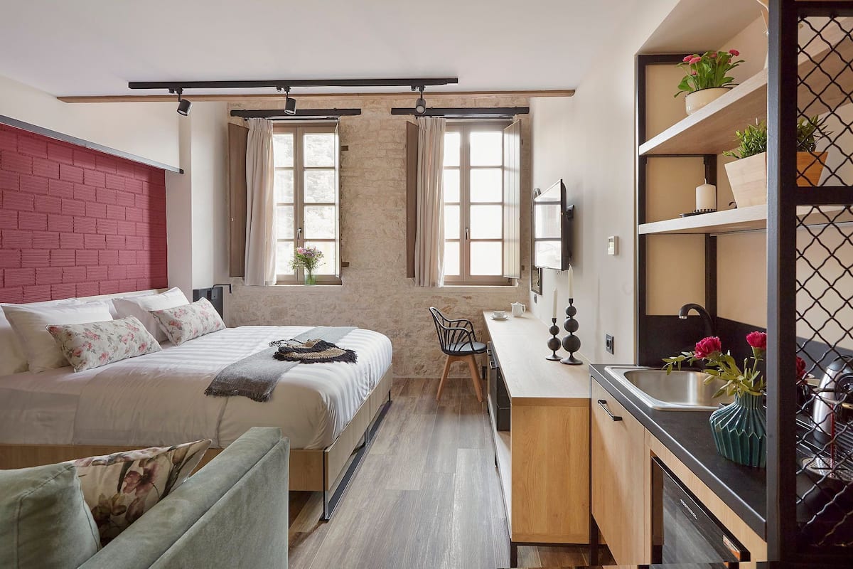 Un dormitorio con cama, escritorio, silla, ventana y una zona de cocina con lavabo y estantes.