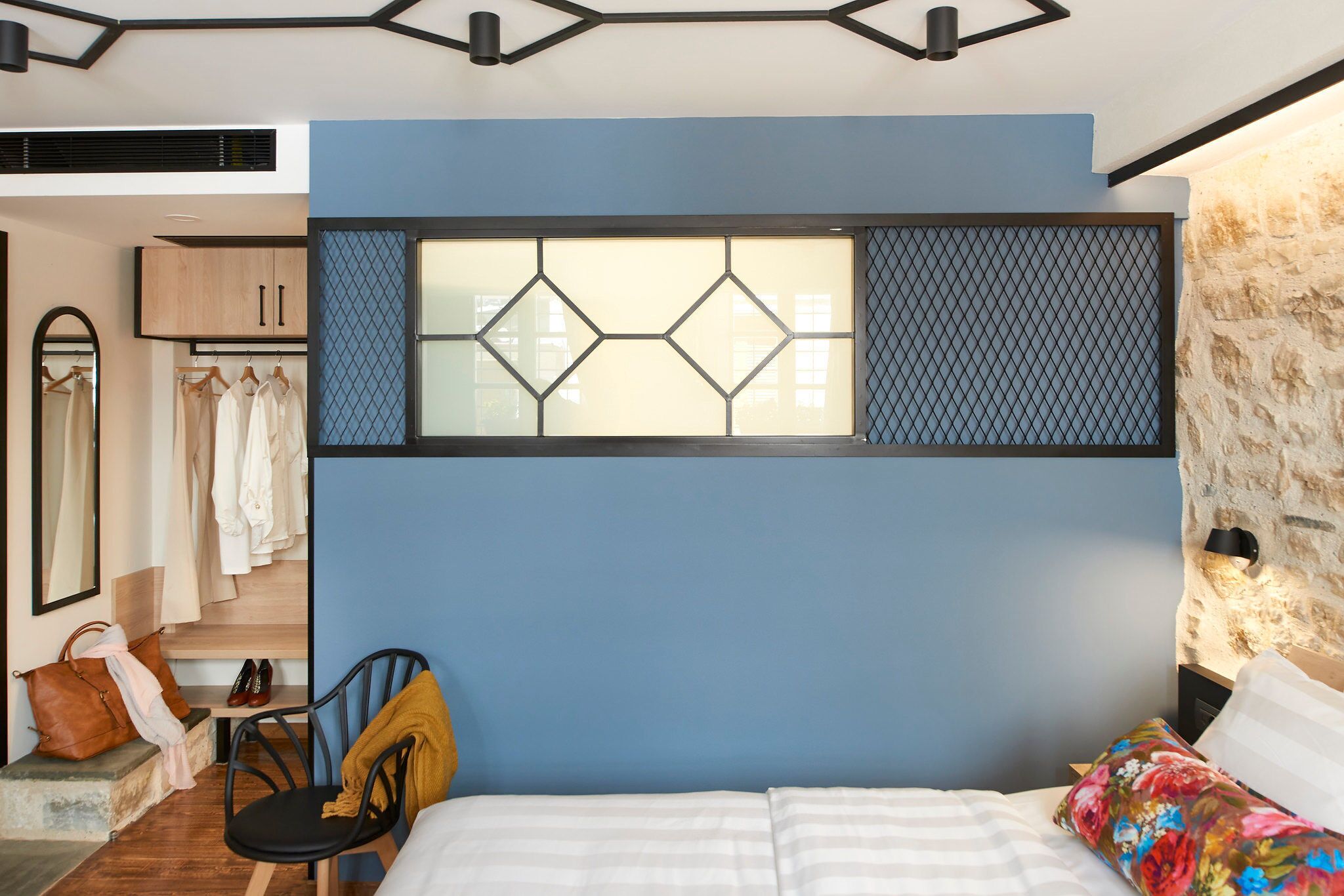 A bedroom with a bed, a chair, a desk with a chair, a wardrobe, and a mirror.