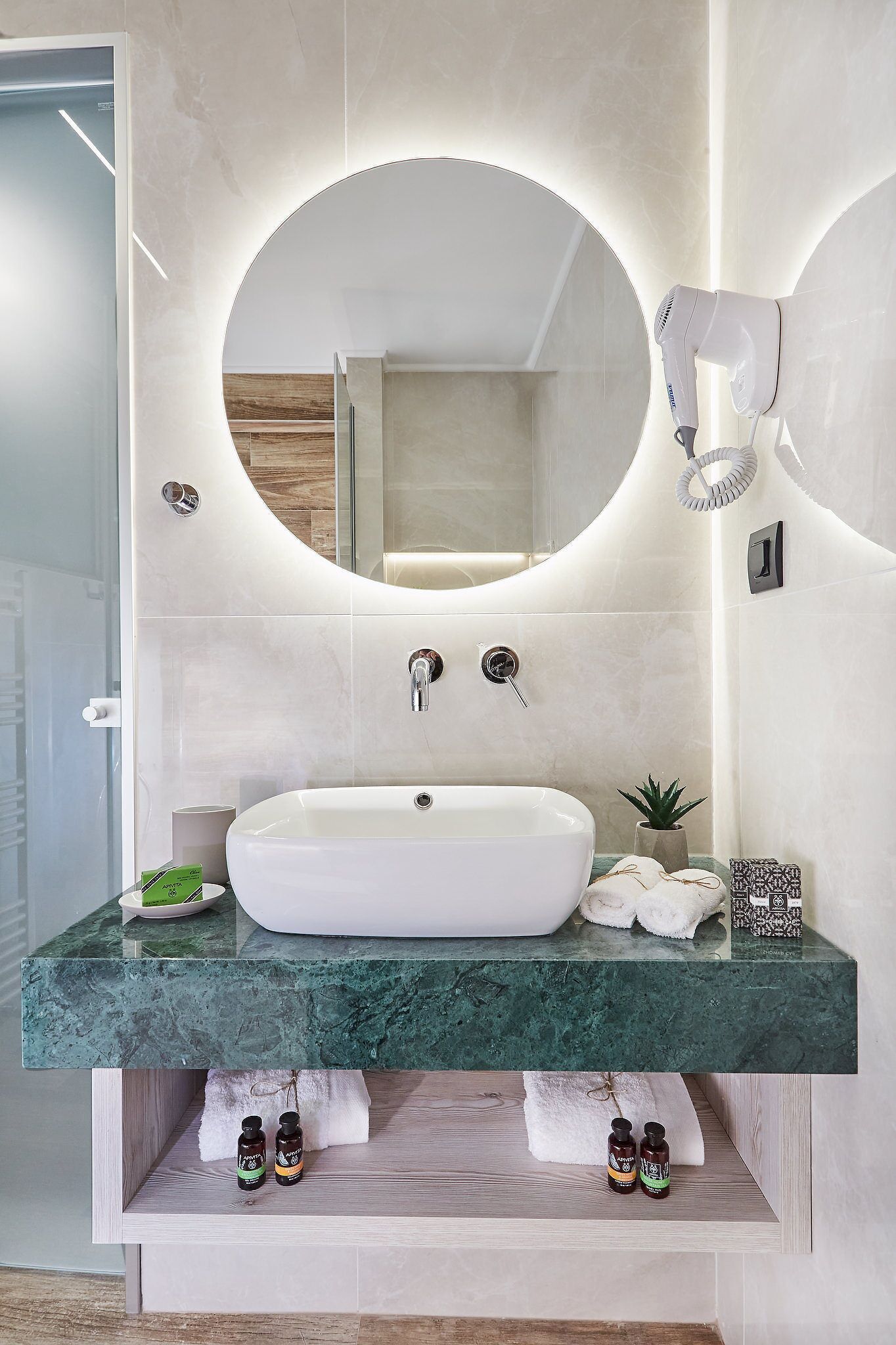 Un baño moderno con un espejo ovalado grande, un lavamanos blanco y un secador de pelo montado en la pared.
