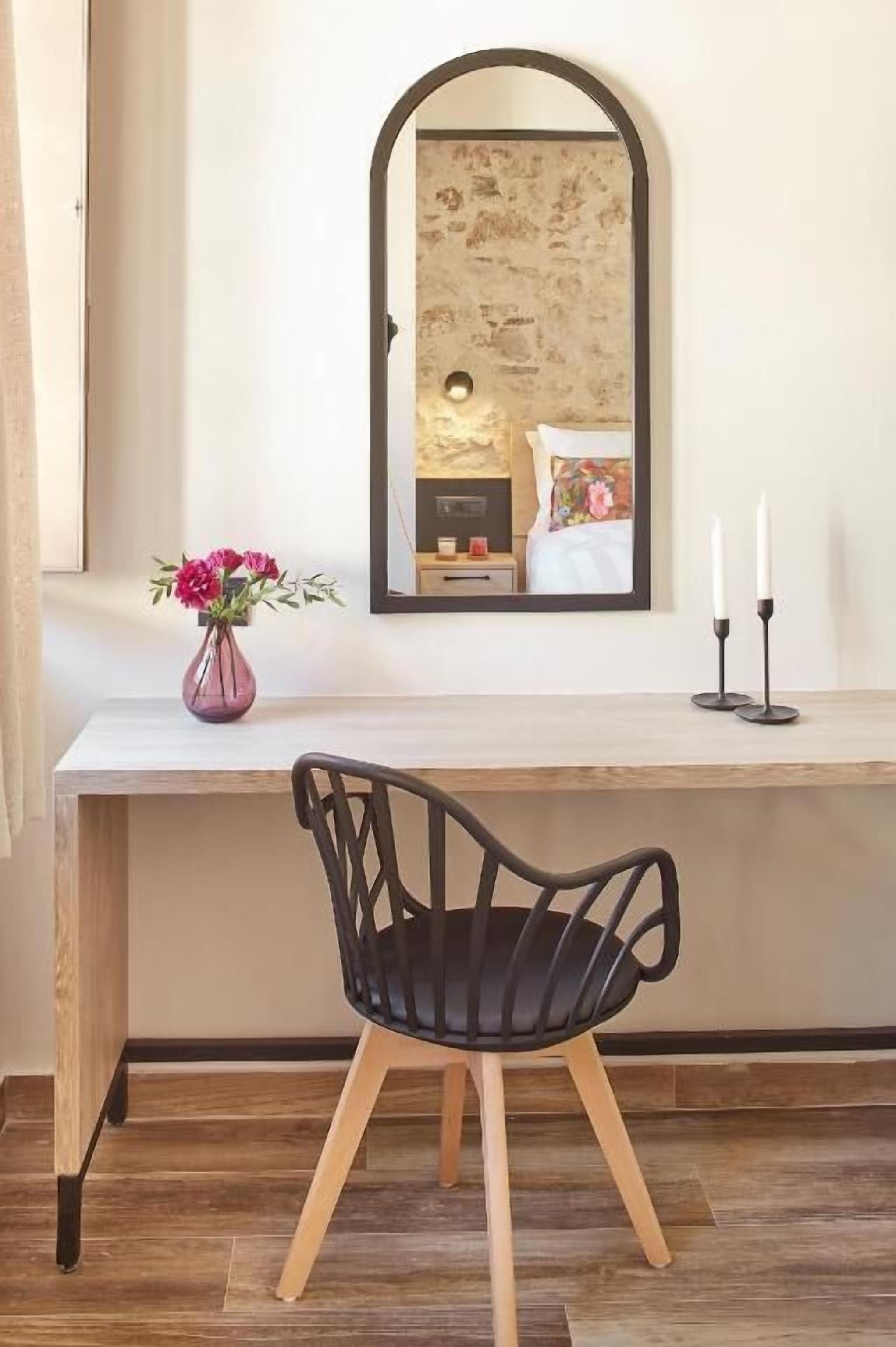 A wooden desk with a black chair, a vase of flowers, and a mirror on the wall.