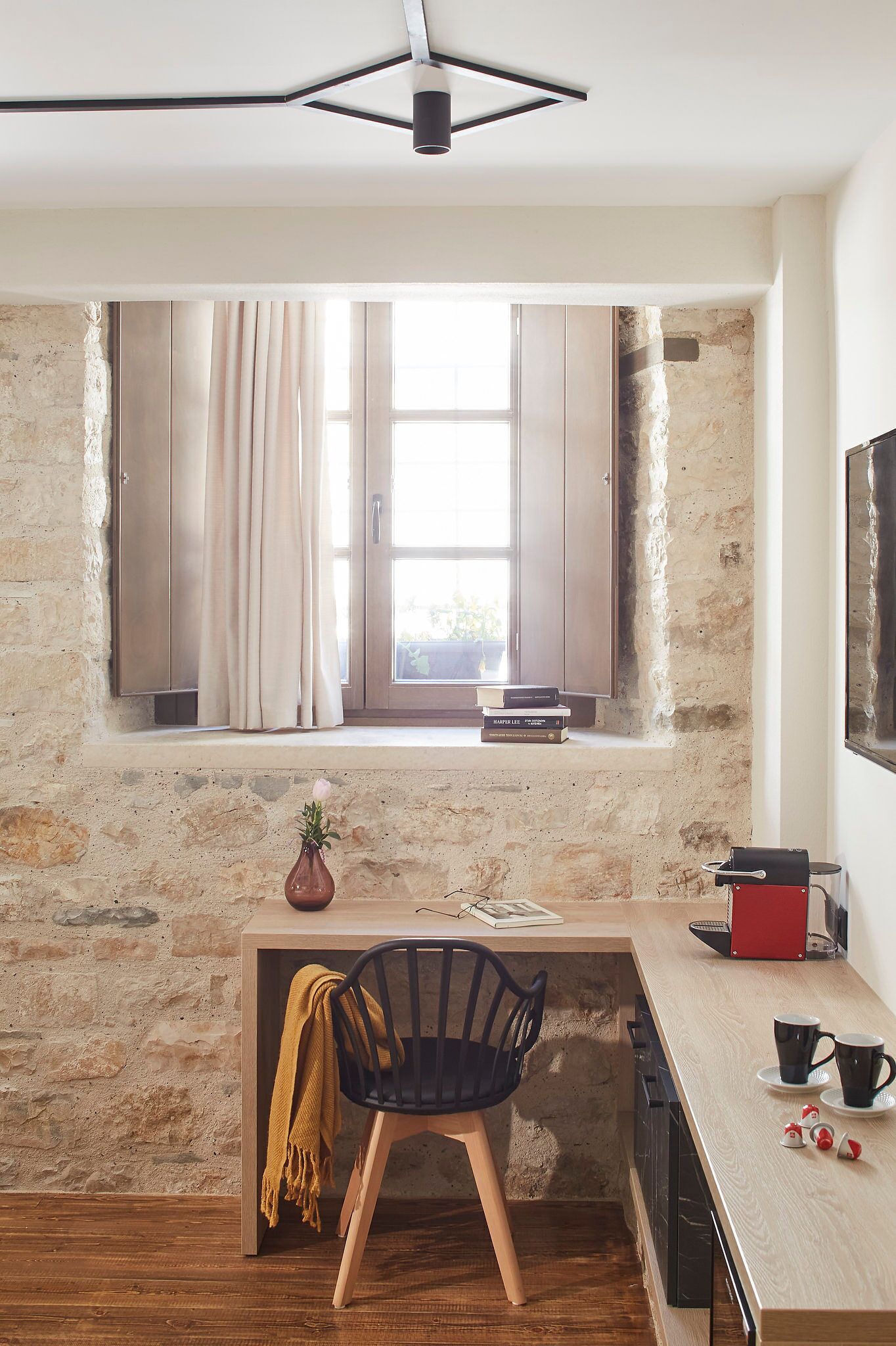 A cozy room with a stone wall, a wooden desk, a chair, a window with curtains, and a television on the wall.