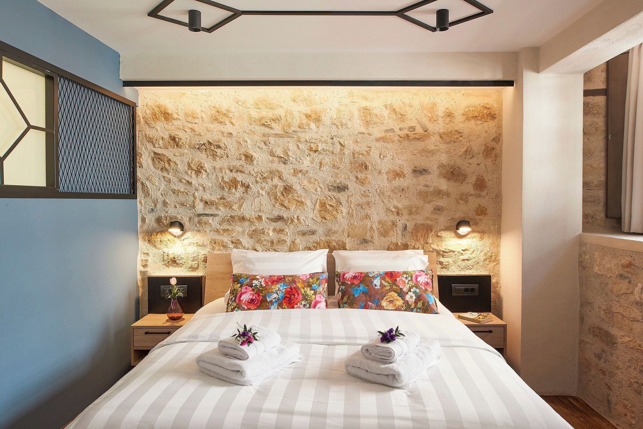 Une chambre avec une tête de lit en pierre, deux tables de chevet avec des lampes, un lit avec une literie à rayures et des coussins à motifs floraux.