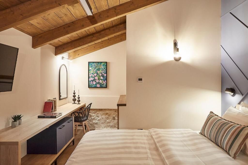 Un dormitorio con techo de madera, una cama con ropa de cama a rayas, un escritorio con silla, un cuadro en la pared y una mesita de noche con lámpara.