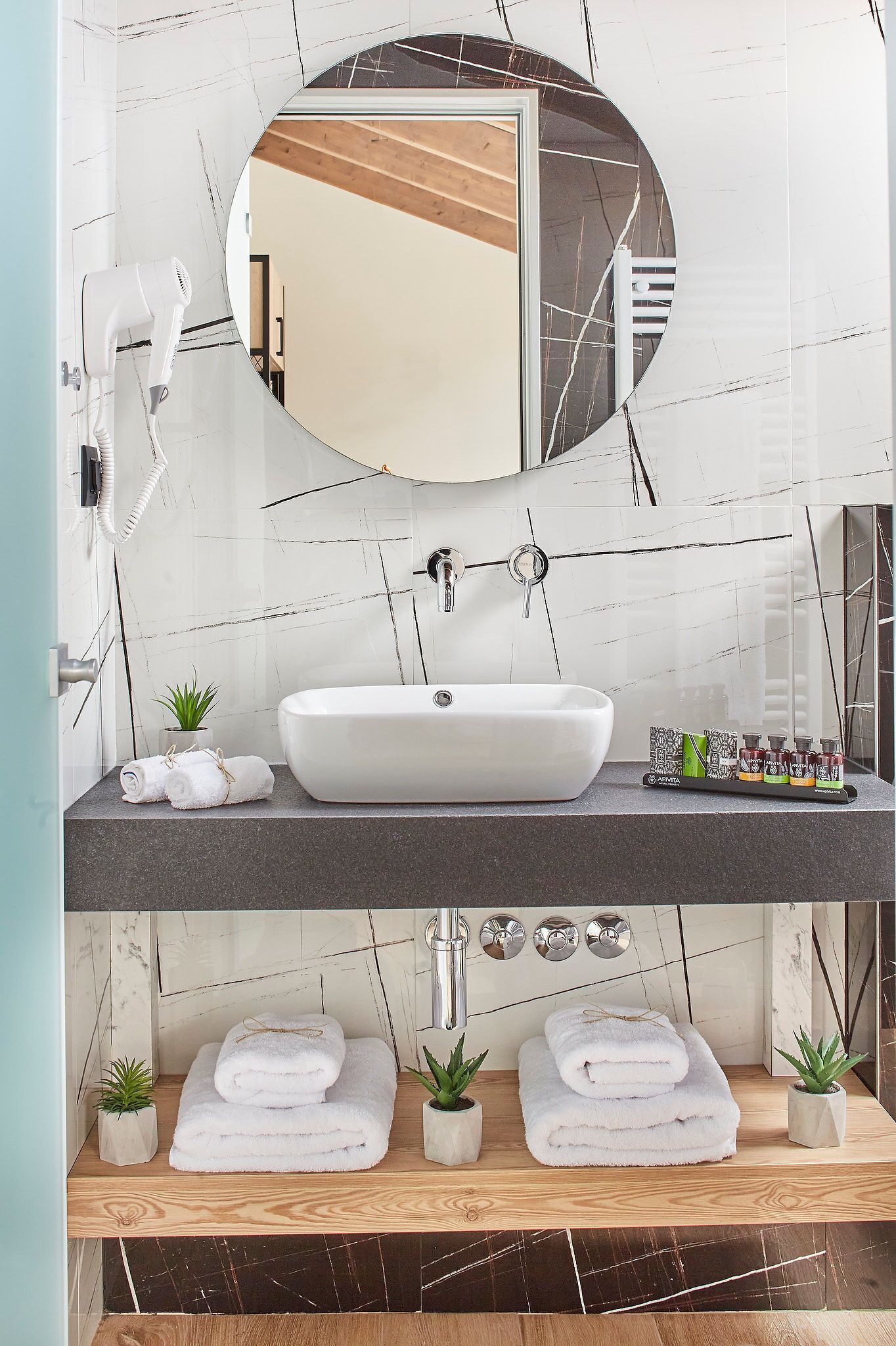 Ein Badezimmer mit einem weißen Waschbecken, einem runden Spiegel und einem an der Wand befestigten Föhn. Auf der Ablage liegen Handtücher und kleine Topfpflanzen.