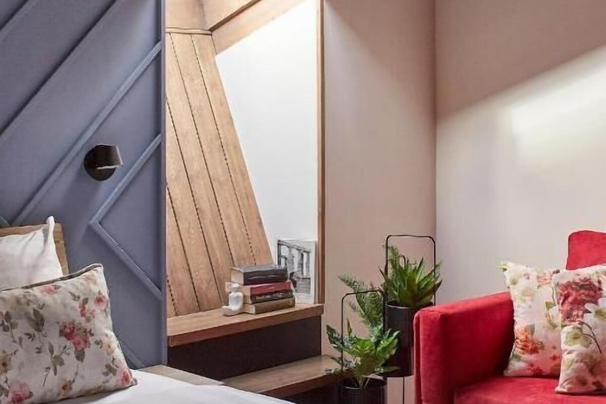 A bedroom with a sloped ceiling, a bed with striped bedding, a red chair, and a small table with books and plants.