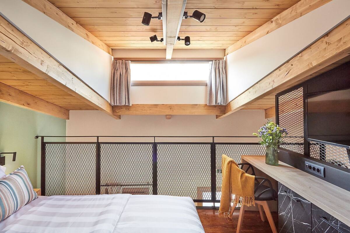 Un dormitorio con cama, escritorio y silla, ventana con cortinas y balcón con barandilla de malla.