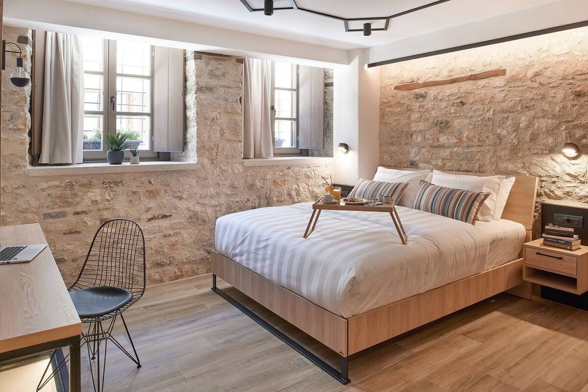 Un dormitorio con una cama de madera, un escritorio con una computadora portátil, una silla y una mesita de noche con una lámpara.