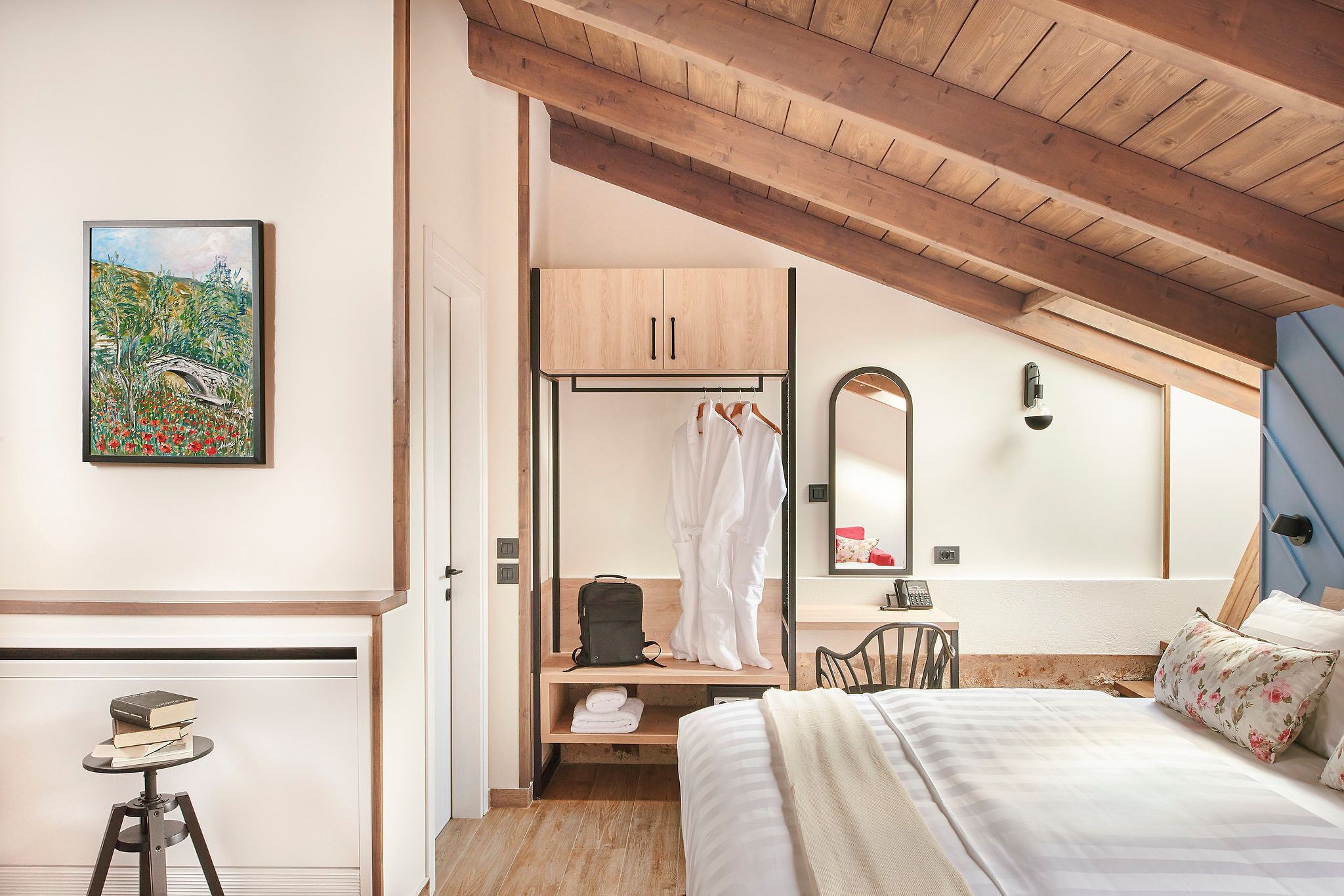 A bedroom with a bed, a wooden ceiling, a painting, a wardrobe, a mirror, and a desk with a chair.
