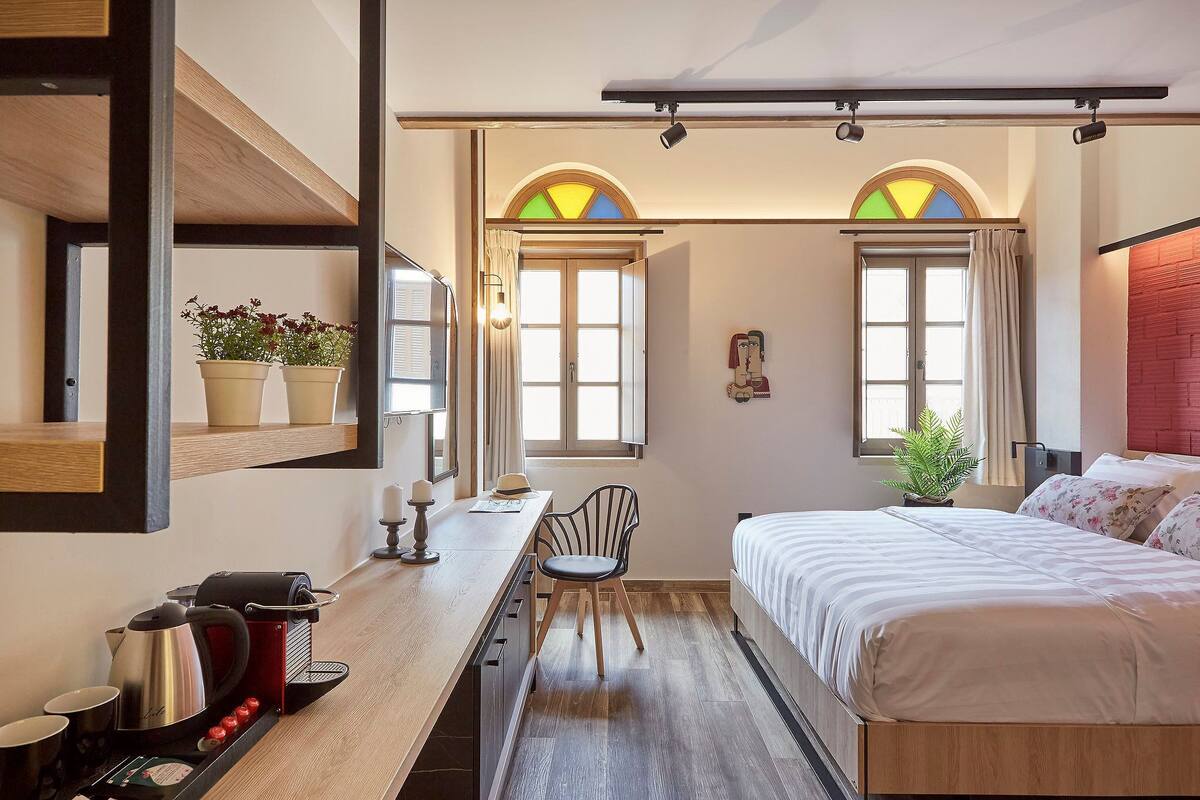 Una habitación de hotel compacta con cama, escritorio con cafetera y dos ventanas con cortinas.