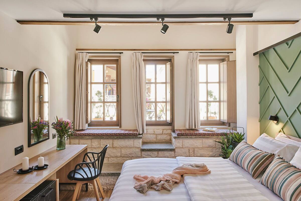 Un dormitorio con cama, mesita de noche, silla y escritorio con espejo. Hay ventanas con cortinas y vistas al exterior.