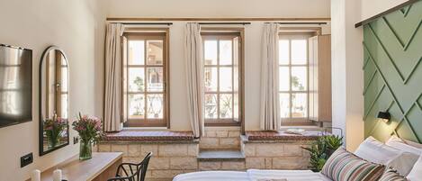 A bedroom with a bed, bedside table, chair, and a desk with a mirror. There are windows with curtains and a view of the outside.