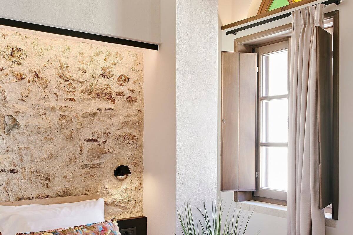 Un dormitorio con pared de piedra, una cama con almohadillas florales, una mesita de noche con una planta y una ventana con cortina.