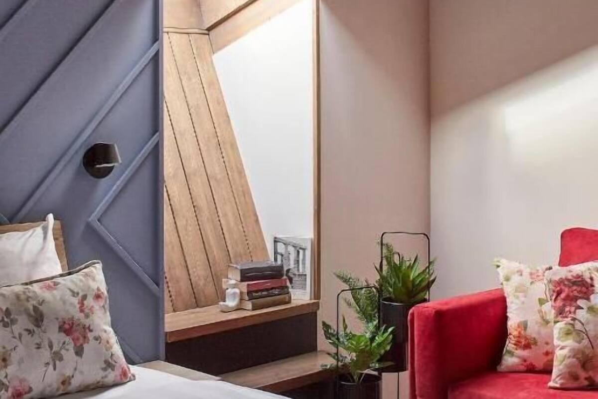 Un dormitorio con techo inclinado, una cama con ropa de cama a rayas, una silla roja y una mesita con libros y plantas.