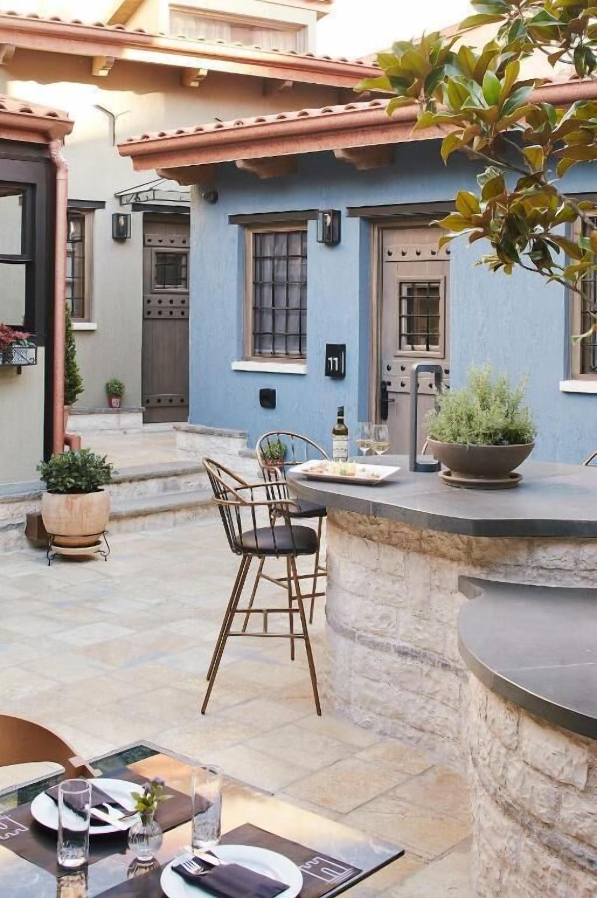 An outdoor dining area with stone tables and chairs, a blue building with a tiled roof in the background, and potted plants.