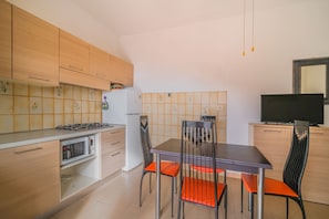 Eine kleine Küche mit Holzausstattung, einem Gasherd, einer Mikrowelle und einem Kühlschrank. Ein Esstisch mit Stühlen und ein Fernseher auf einem Schrank sind ebenfalls vorhanden.