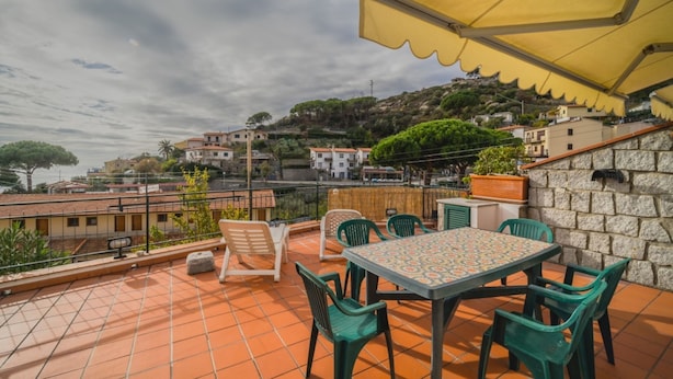 A terrace with a table and chairs, a yellow umbrella, and a view of a hillside with buildings. - LA TERRAZZA A SECCHETO SUL MARE (Campo nell'Elba)