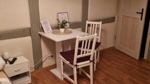 A small room with a wooden floor, a white desk with a chair, a small bedside table with a lamp, and a door. - Room 08 (DH) - Pension Torgau (Torgau)