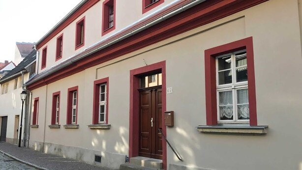 A building with a red door and windows, a cobblestone street, and a small set of steps leading to the entrance. - Room 08 (DH) - Pension Torgau (Torgau)