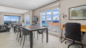 Una stanza spaziosa con un tavolo grande, sedie, una scrivania con un computer e una sedia con rotelle. È presente un'ampia finestra vista mare e una porta che dà su un'altra stanza.