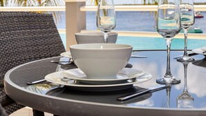 Tavola apparecchiata con piatti e bicchieri bianchi, con vista su una piscina, una palma e una barca sullo sfondo.