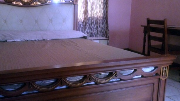 A wooden bed with a headboard and drawers, a chair, and a window with patterned curtains. - Sooper Guest House (Kisumu)