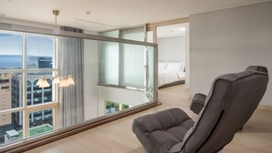 Une chambre d’hôtel moderne avec une grande fenêtre, un fauteuil gris, un lit et une vue sur le paysage urbain.