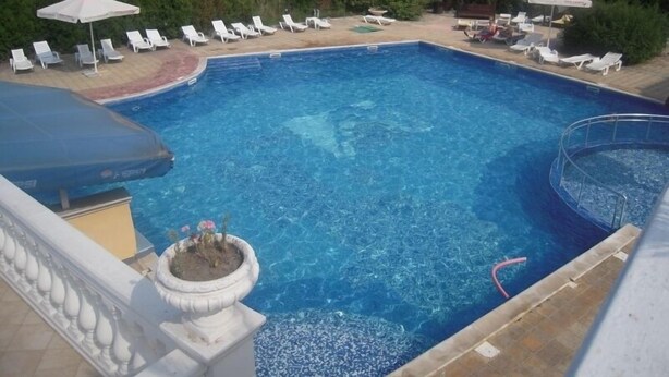 Una piscina rectangular con agua azul cristalina, rodeada de sillas de descanso y sombrillas blancas, con una barandilla y una maceta en primer plano.