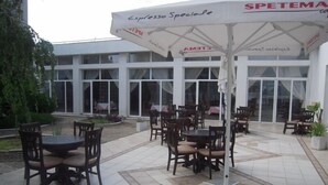 Un área de patio al aire libre con varias mesas y sillas, un paraguas grande y un edificio con ventanas amplias al fondo.
