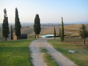 Un sentiero sterrato conduce a una zona piscina circondata da cipressi, con un piccolo edificio in legno e una vista aperta su dolci colline sullo sfondo.