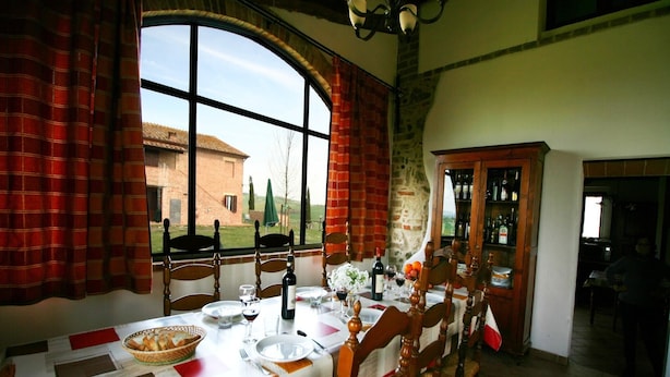 A dining area with a long table set for six, wooden chairs, a chandelier, and a view of a brick building outside. - Charming Tuscan Farmhouse (Arbia Asciano)