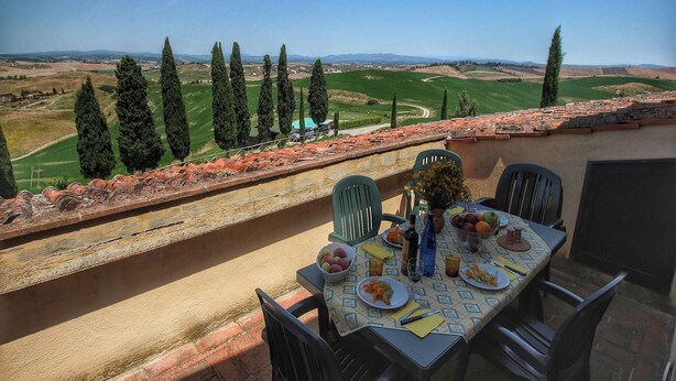 Un área de comedor al aire libre con una mesa dispuesta para dos, con vistas a un paisaje pintoresco de colinas ondulantes y cipreses.