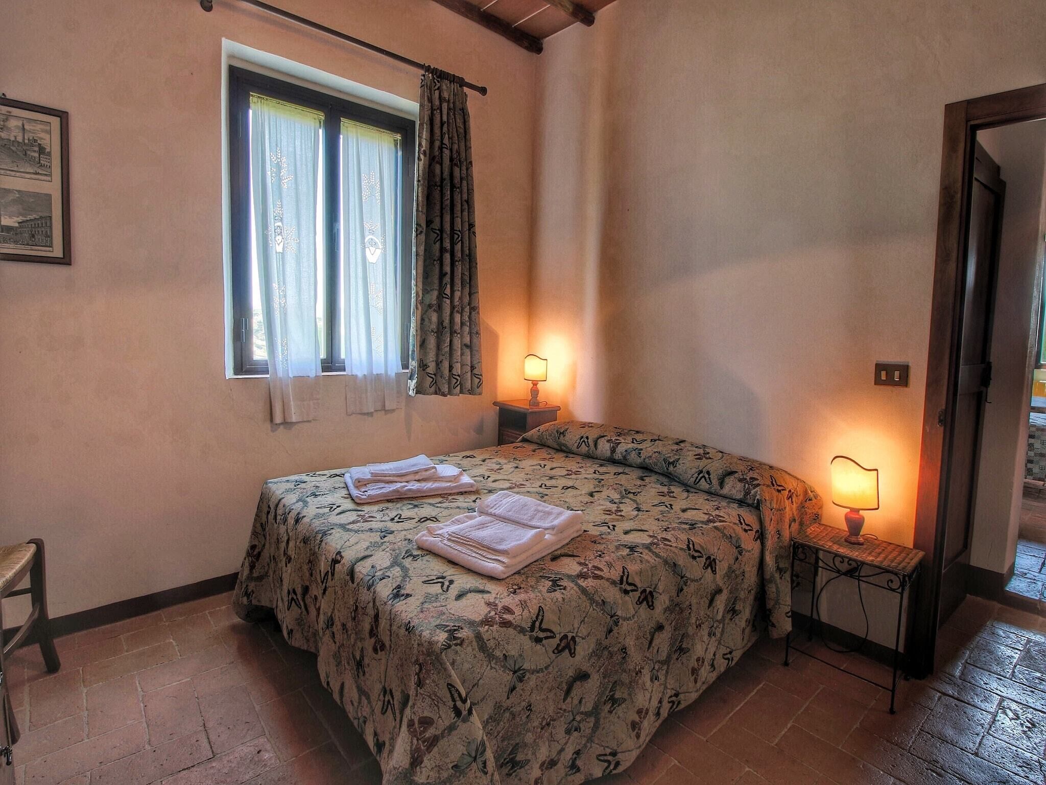 A bedroom with a bed, two bedside lamps, a window with curtains, a chair, and a small table with a lamp.
