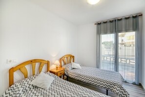 A room with two beds, wooden headboards, a nightstand with a lamp, and a large window with curtains.