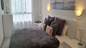 Et soveværelse med seng, sengeborde, lamper, en stol, et skrivebord og et vindue med gardiner.