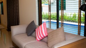 A modern living room with a beige sofa, two pillows, a wooden headboard, and a view of a pool through large windows. - Hua-Hin Private pool villa (Tambon Hin Lek Fai, Amper Hua-Hin)