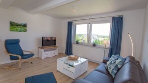 A modern living room with a sofa, a blue armchair, a glass coffee table, a TV, and a window with curtains. - 5 star FeWo Boddenblick (Glowe)