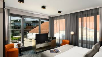 A modern hotel room with a large bed, a television, a desk with a coffee machine, and a view of the city through large windows.