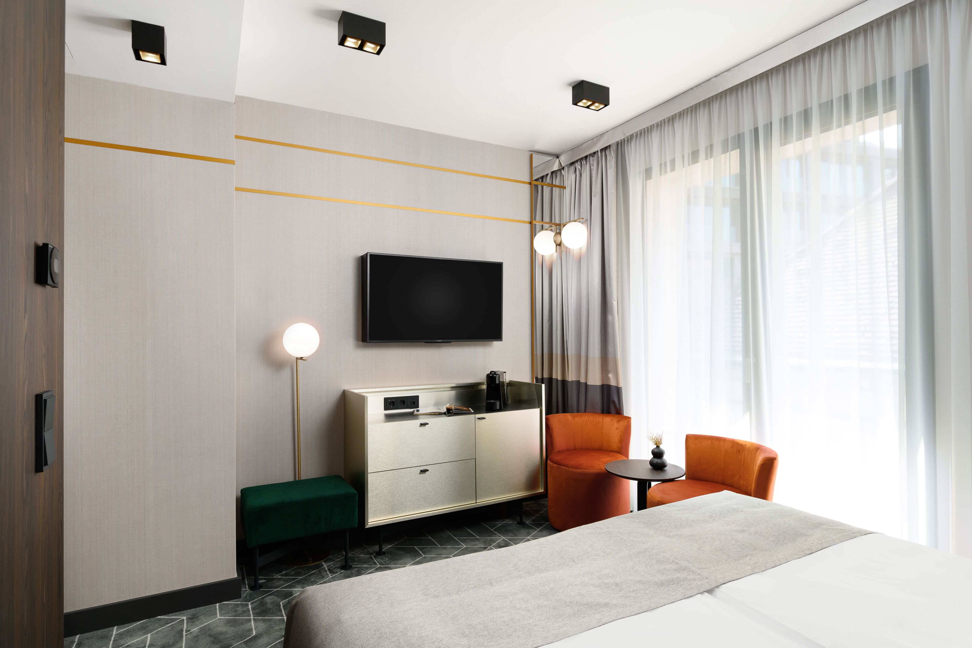 A modern hotel room with a large bed, a TV mounted on the wall, a seating area with a small table, and a large window with curtains.