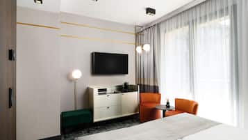 A modern hotel room with a large bed, a TV mounted on the wall, a seating area with a small table, and a large window with curtains.