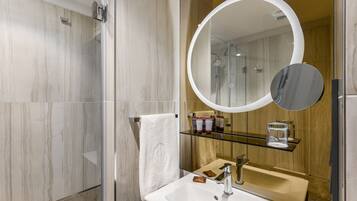 Un baño moderno con cabina de ducha de vidrio, un lavabo blanco, un espejo redondo y un mueble de tocador de madera.