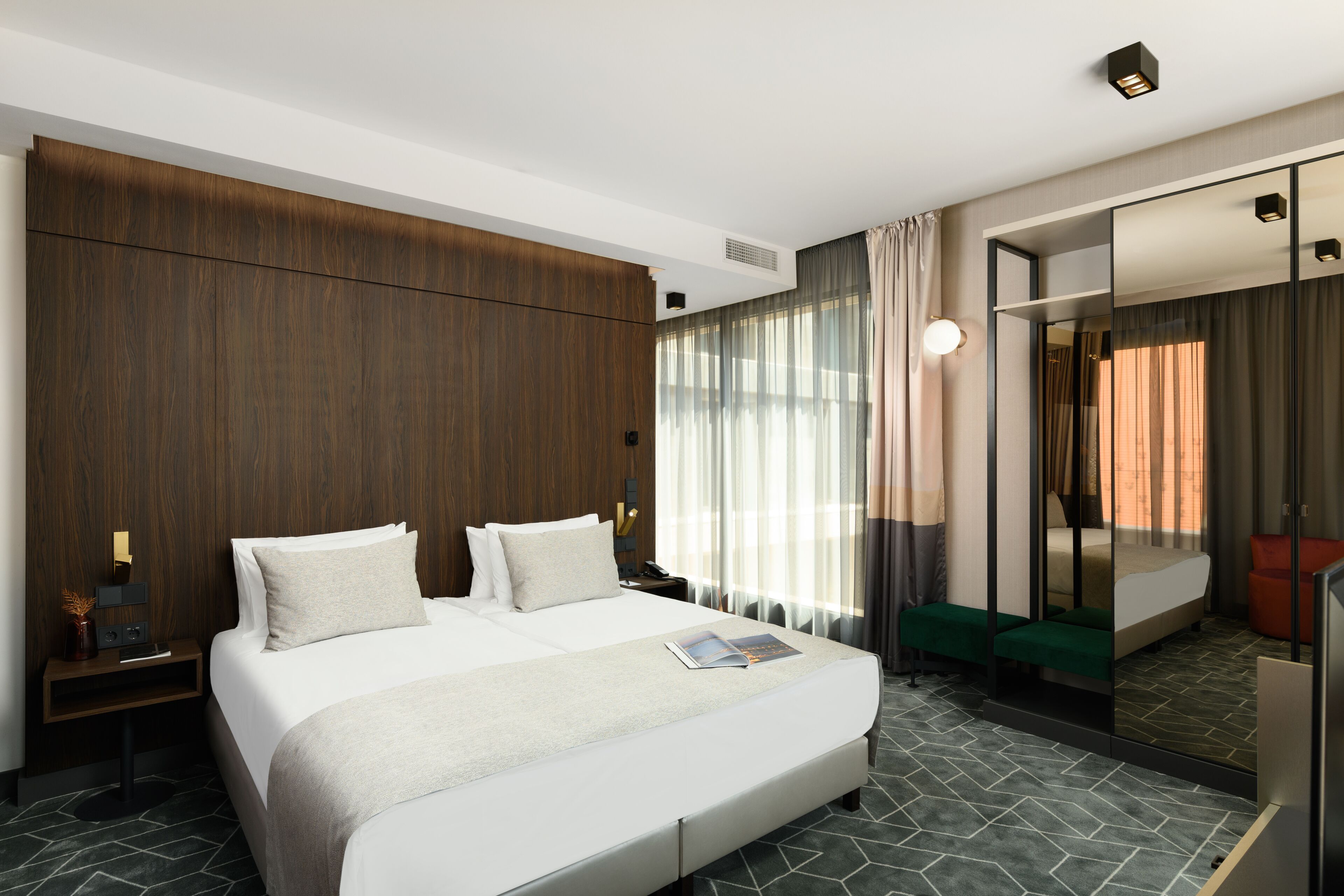 Una camera d'albergo moderna con un letto grande, un comodino, uno specchio e vista sulla città attraverso ampie finestre.
