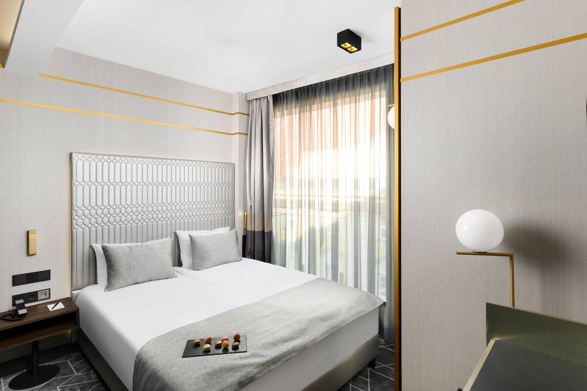A modern hotel room with a large bed, a bedside table with a lamp, and a view of the outdoors through sheer curtains.