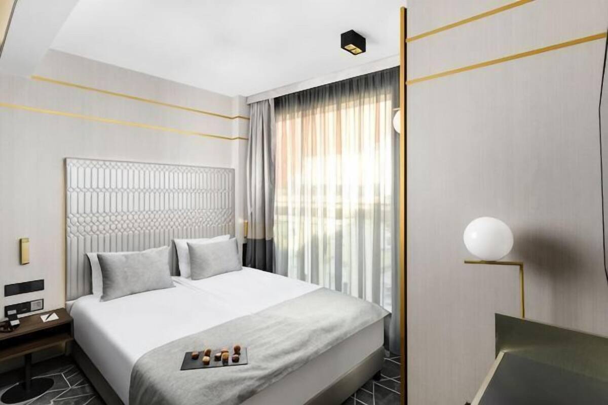 A modern hotel room with a large bed, a bedside table, a lamp, and a view of the outdoors.