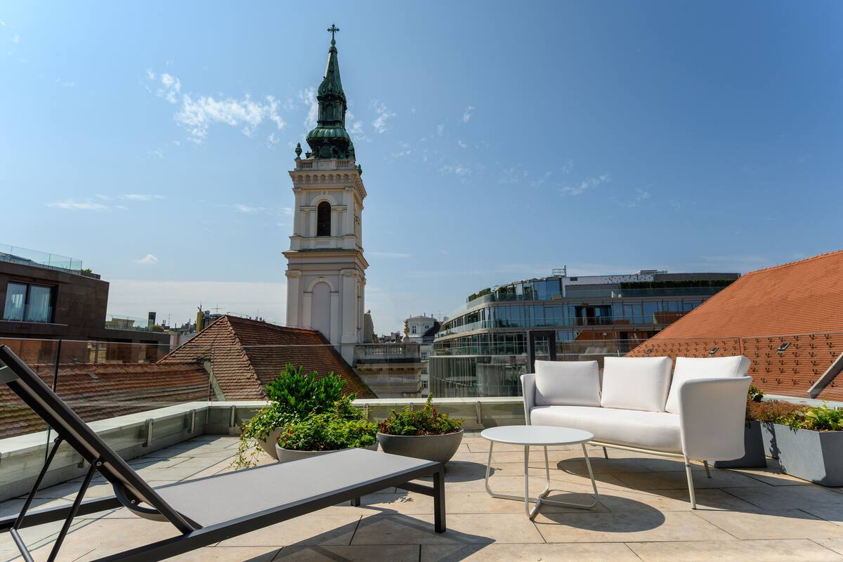 A rooftop terrace with a white sofa, a small table, and potted plants. A church steeple is visible in the background.